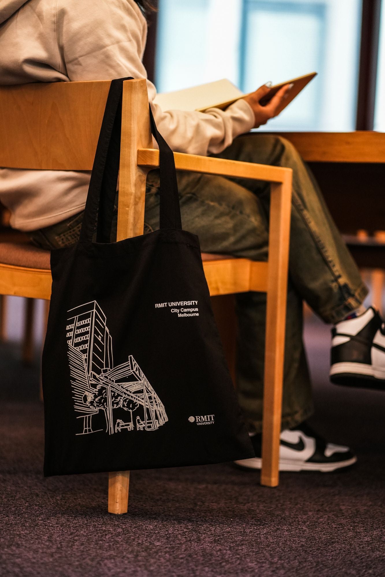 Black Limited Edition Campus Tote with RMIT University City Campus design, hanging on the backrest of a light wooden chair beside a seated model wearing a beige hoodie, green jeans, and black-and-white sneakers while holding an open notebook. The tote features a white architectural line-art design of a Melbourne campus building with trees and beams, and includes two white printed logos: “RMIT UNIVERSITY City Campus Melbourne” and the RMIT logo. Captured in an indoor study environment.